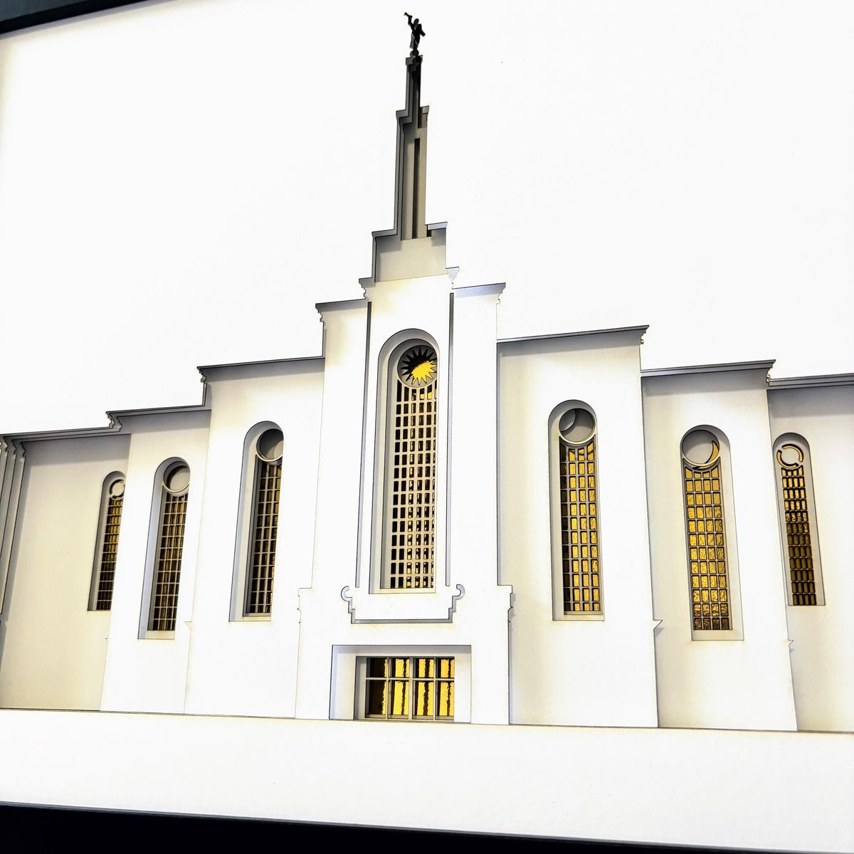 A white architectural model or miniature building facade with tall arched windows featuring gold-toned gridded panes, a central spire with a small statue on top, and a bright white background.