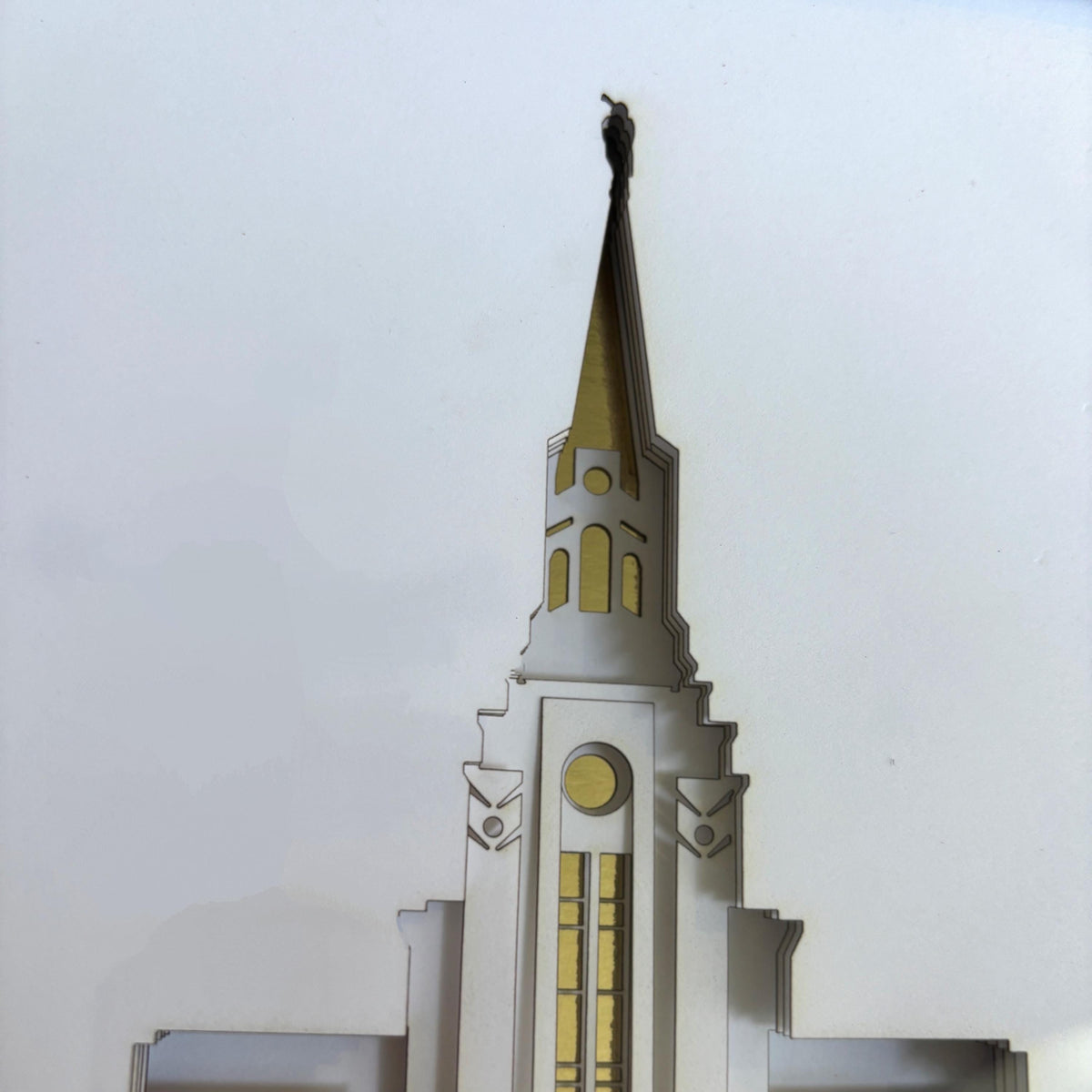 A close-up of a white and gold architectural spire design, likely a decorative wall art piece or model, set against a plain light gray background.