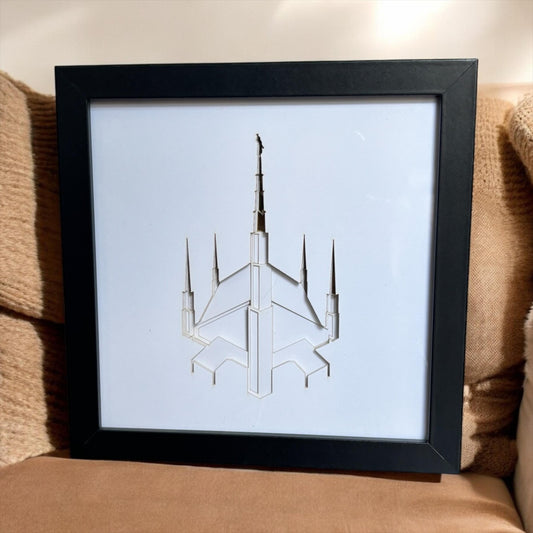A framed art print featuring a minimalist white line drawing of a futuristic spacecraft or spire, set in a black frame and displayed on a brown textured couch. Dallas Temple Art Gift.