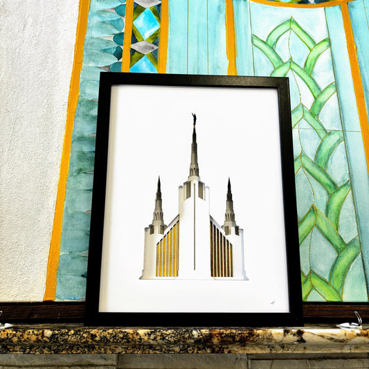 A framed art print featuring a stylized white temple illustration with gold accents, displayed on a stone ledge in front of a colorful stained-glass-style wall.