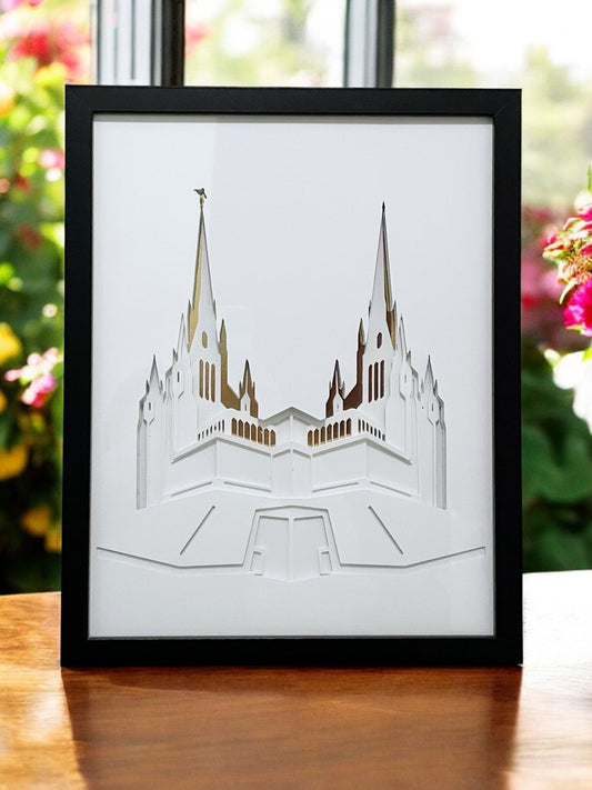 A framed laser-cut architectural art print of the Salt Lake Temple, featuring white layered cutouts with gold foil highlights inside a black frame, displayed on a wooden table with a blurred garden background. San Diego Temple Art Shadow Box