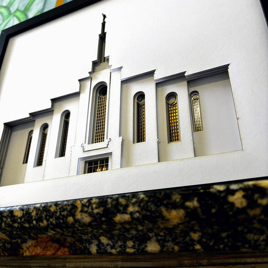 A miniature architectural model of a building facade with arched windows and a central spire, mounted on a white backing and photographed at an angle above a dark marbled surface.