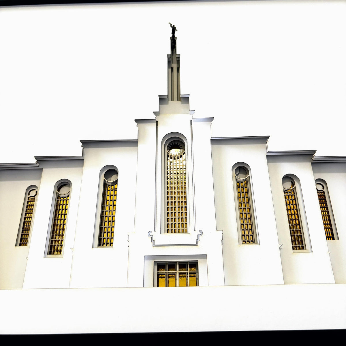 Front-facing view of a white temple building with tall arched windows featuring yellow-tinted glass and a central spire topped with a statue, shown against a bright sky.