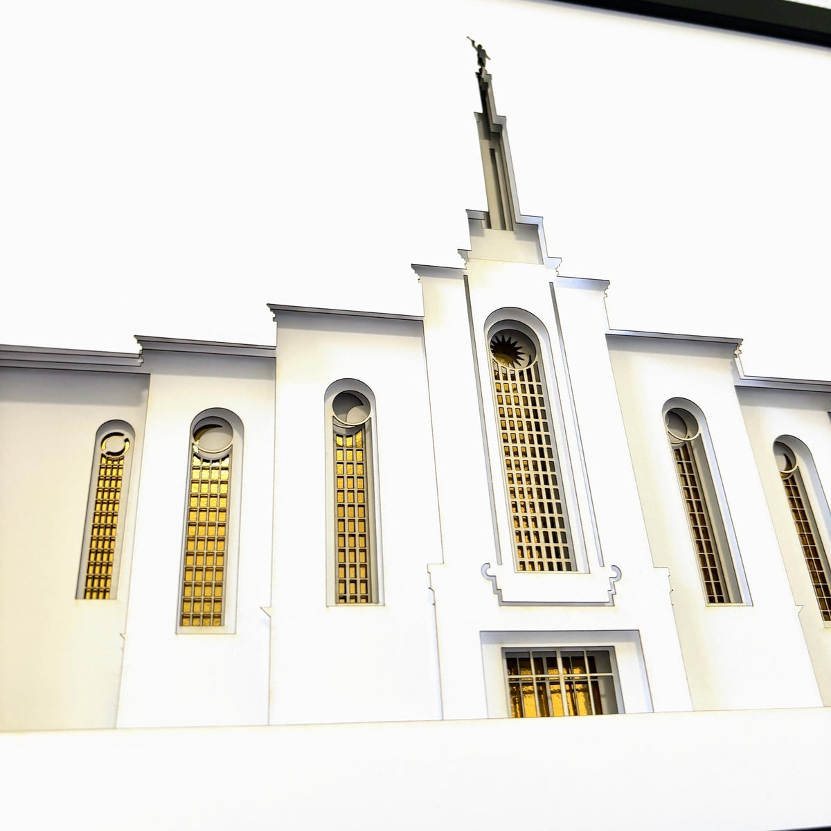 A low-angle photograph of a white architectural building facade with tall arched windows featuring gold grid patterns and a spire topped with a statue Albuquerque Temple Art