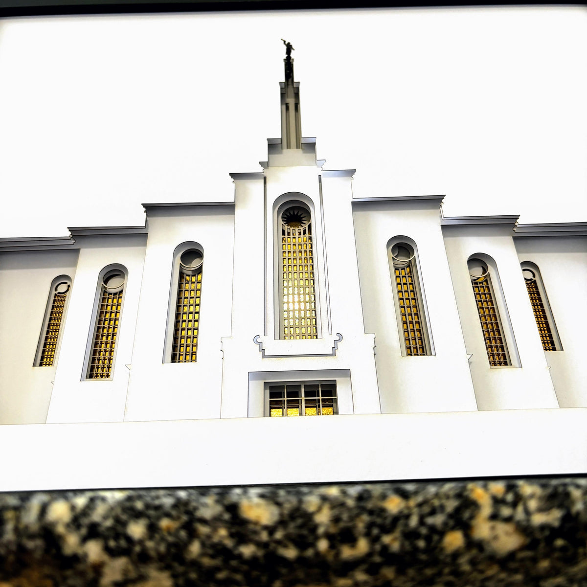 A low-angle photograph of a white Albuquerque temple building with gold-toned arched windows and a statue atop its spire, set against a bright white sky.