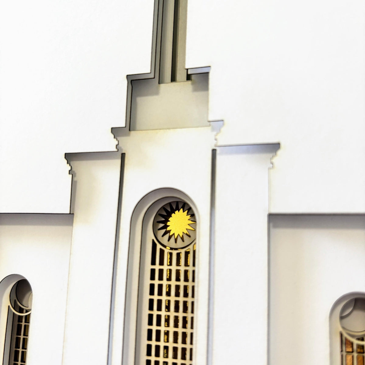 A close-up of a white architectural model or miniature featuring a central arched window with a gold sunburst emblem above a grid pattern.