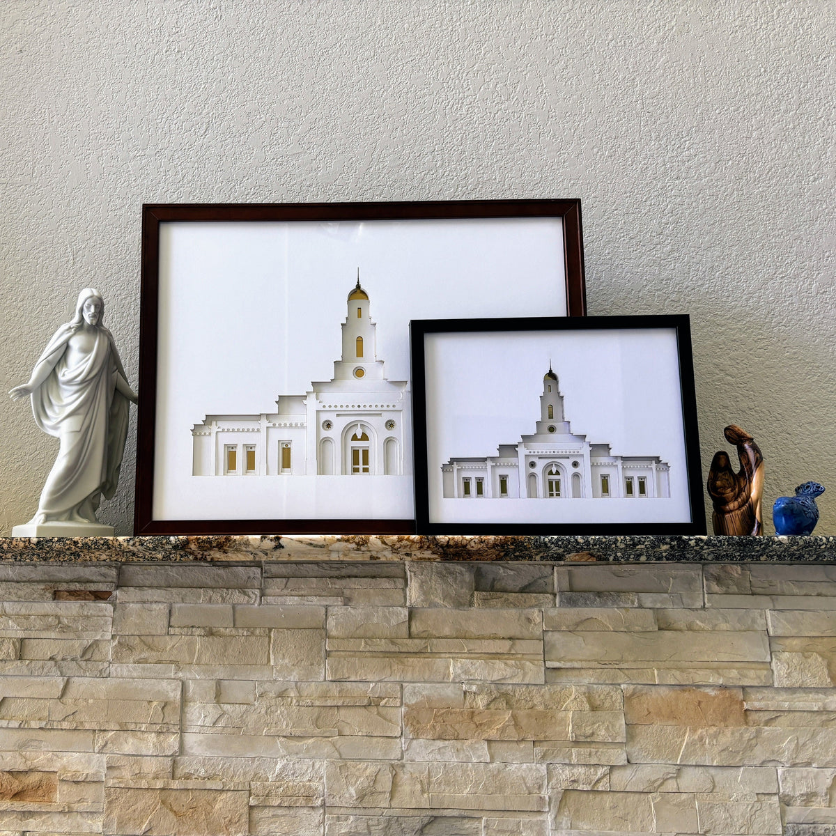 Two framed architectural prints of a white church building, one in a dark brown frame and one in a black frame, displayed on a stone mantel alongside decorative figurines.