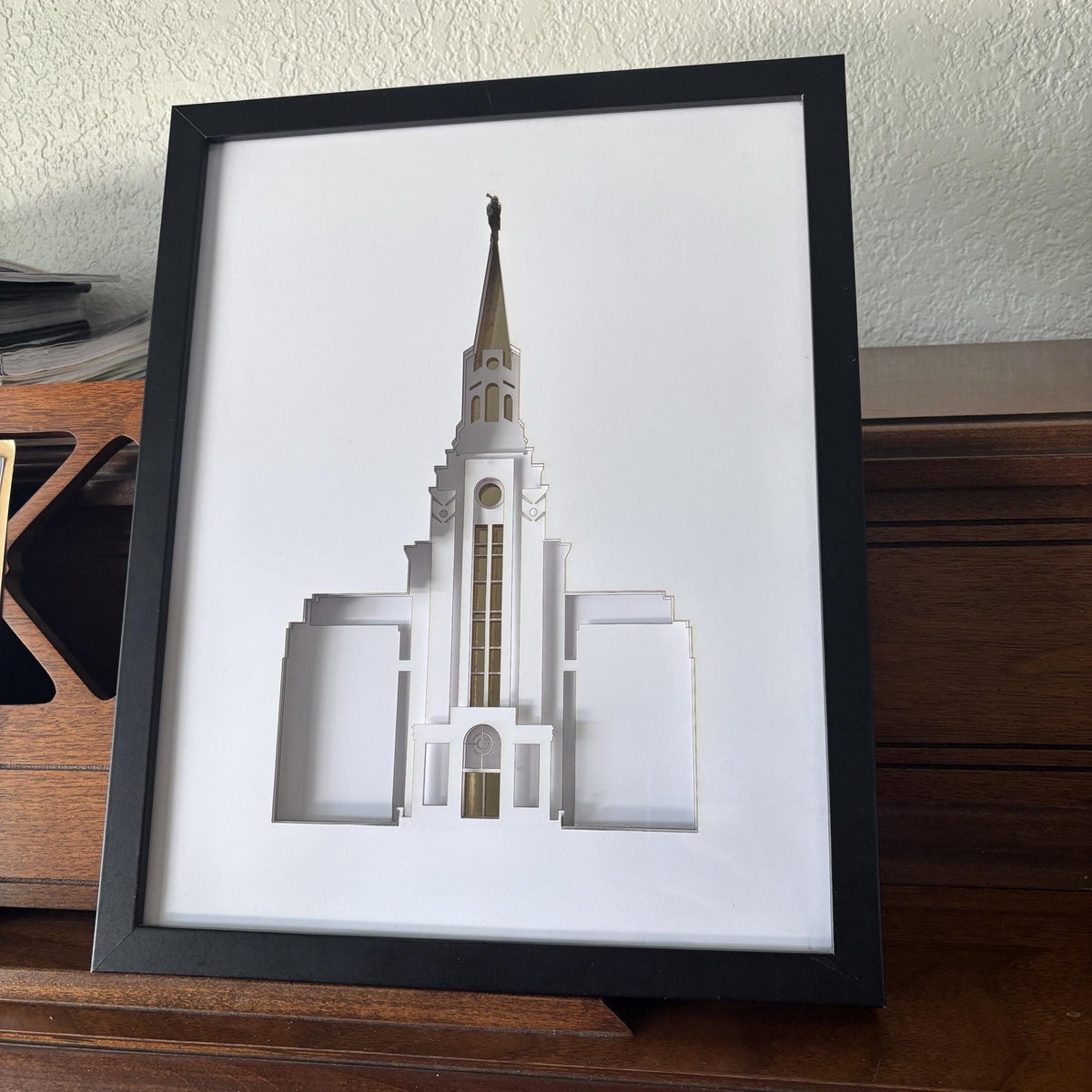 A framed 3D papercut model of a church building with a tall spire, displayed on a wooden surface against a textured wall.