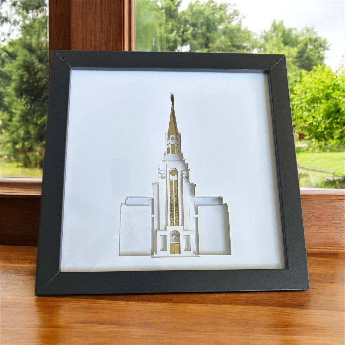 A framed art print featuring a 3D layered architectural model of a white temple with a gold spire, displayed on a wooden surface in front of a window with green trees outside.