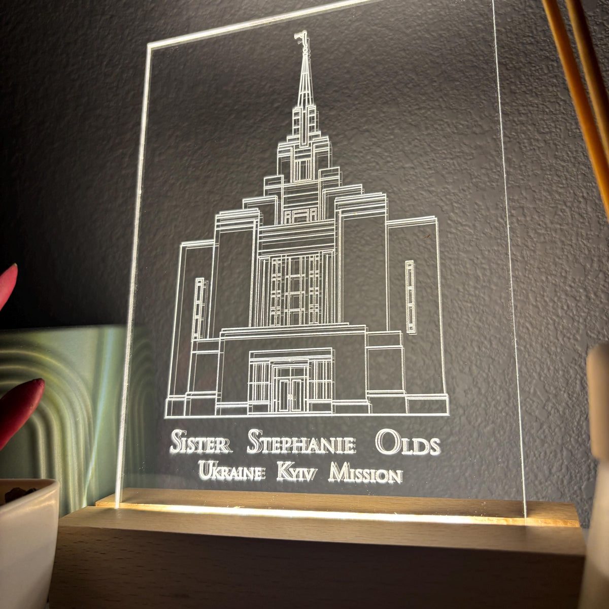 A rectangular acrylic LED night light displaying a white line drawing of a temple building with the engraved text 'Sister. Stephanie Olds Ukraine Ktv Mission' on a wooden base, shown lit against a dark textured wall.