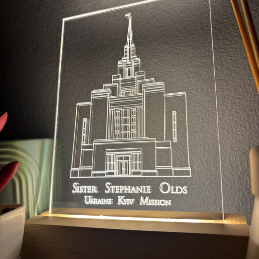 A rectangular acrylic LED night light displaying a white line drawing of a temple building with the engraved text 'Sister. Stephanie Olds Ukraine Ktv Mission' on a wooden base, shown lit against a dark textured wall.