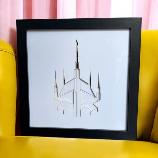 A framed art print featuring a minimalist white line drawing of a futuristic spire-like structure on a white background, set in a black frame and displayed on a yellow chair with pink curtains behind it. Dallas Temple Art