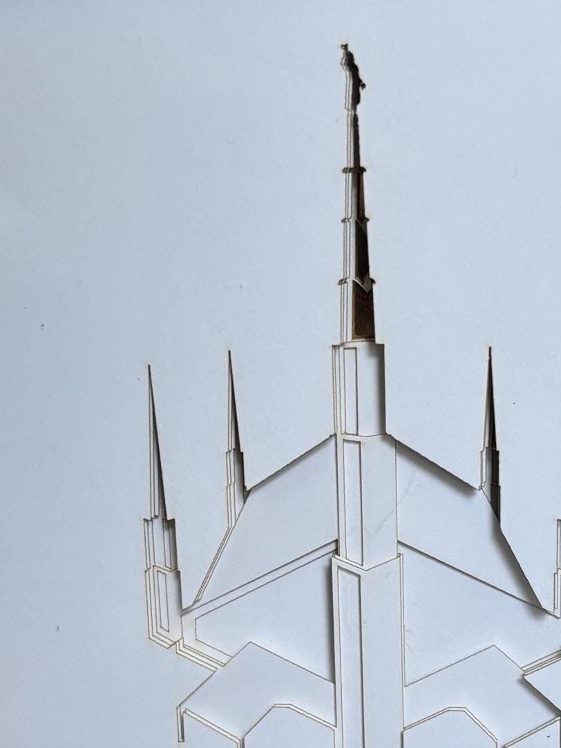 A close-up of a white geometric architectural cutout or sculpture featuring multiple pointed spires and a central tall spire topped with a small bird figure, set against a plain light gray background. Dallas Temple Art LDS