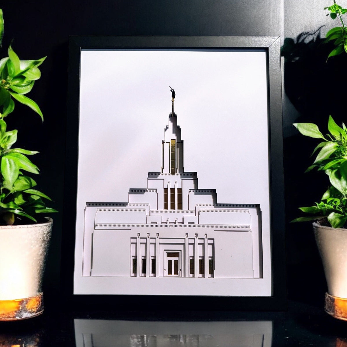 A framed architectural art print of the Salt Lake Temple, displayed on a reflective black surface between two potted green plants.