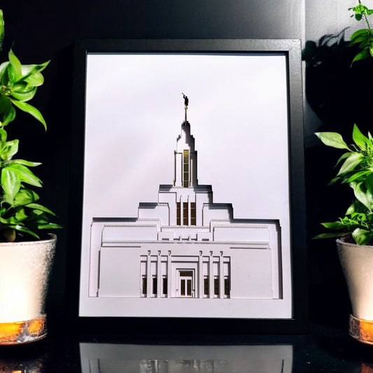 A framed architectural art print of the Salt Lake Temple, displayed on a reflective black surface between two potted green plants.