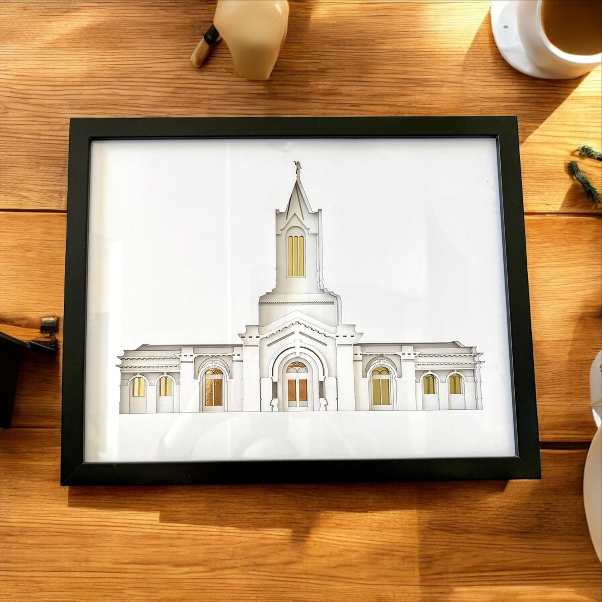A framed architectural art print of a white temple building with gold window accents, displayed on a wooden table next to a coffee cup. Fort Collins Temple Art Gift