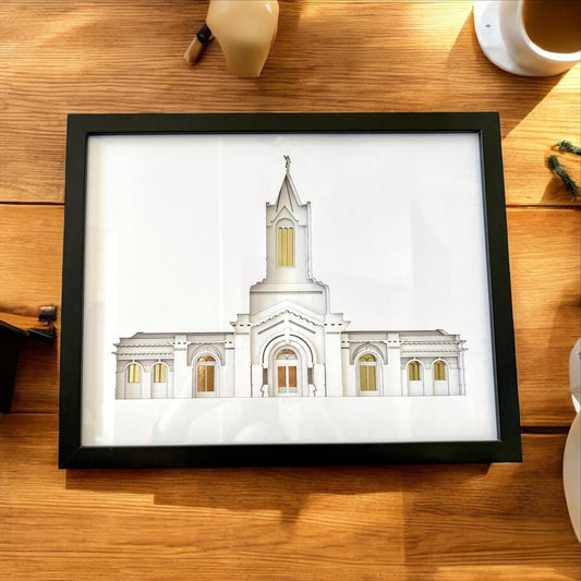 A framed architectural art print of a white temple building with gold window accents, displayed on a wooden table next to a coffee cup. Fort Collins Temple Art Gift