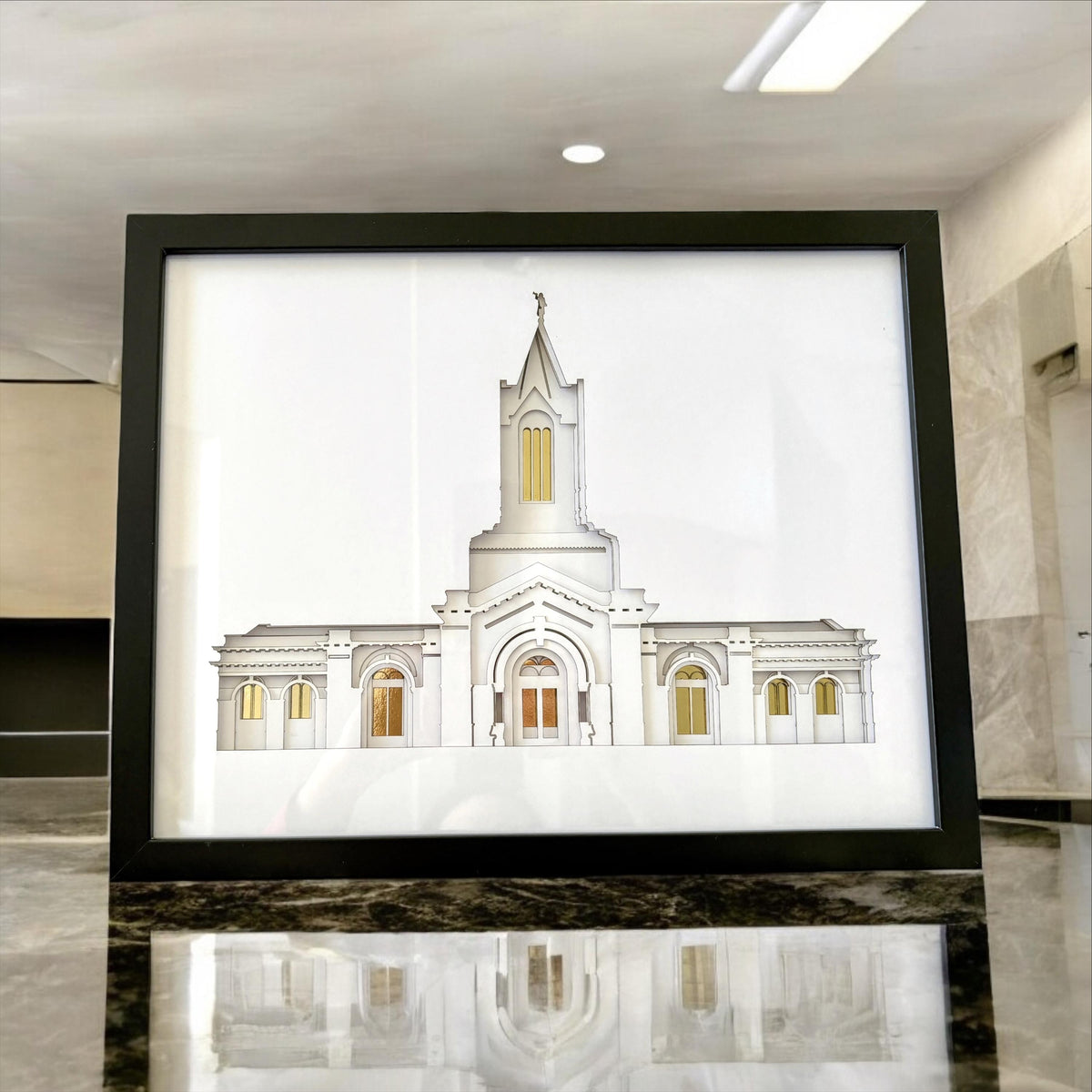 A framed architectural art print of a white temple building with gold window accents, displayed on a polished dark marble surface in a modern interior setting.