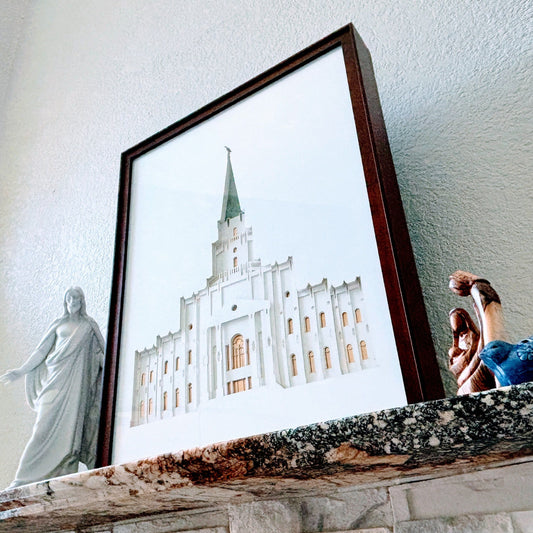A framed art print of a white temple building with a green spire, leaning on a stone mantel next to a white statue and a small figurine, against a textured white wall. Houston Temple Art