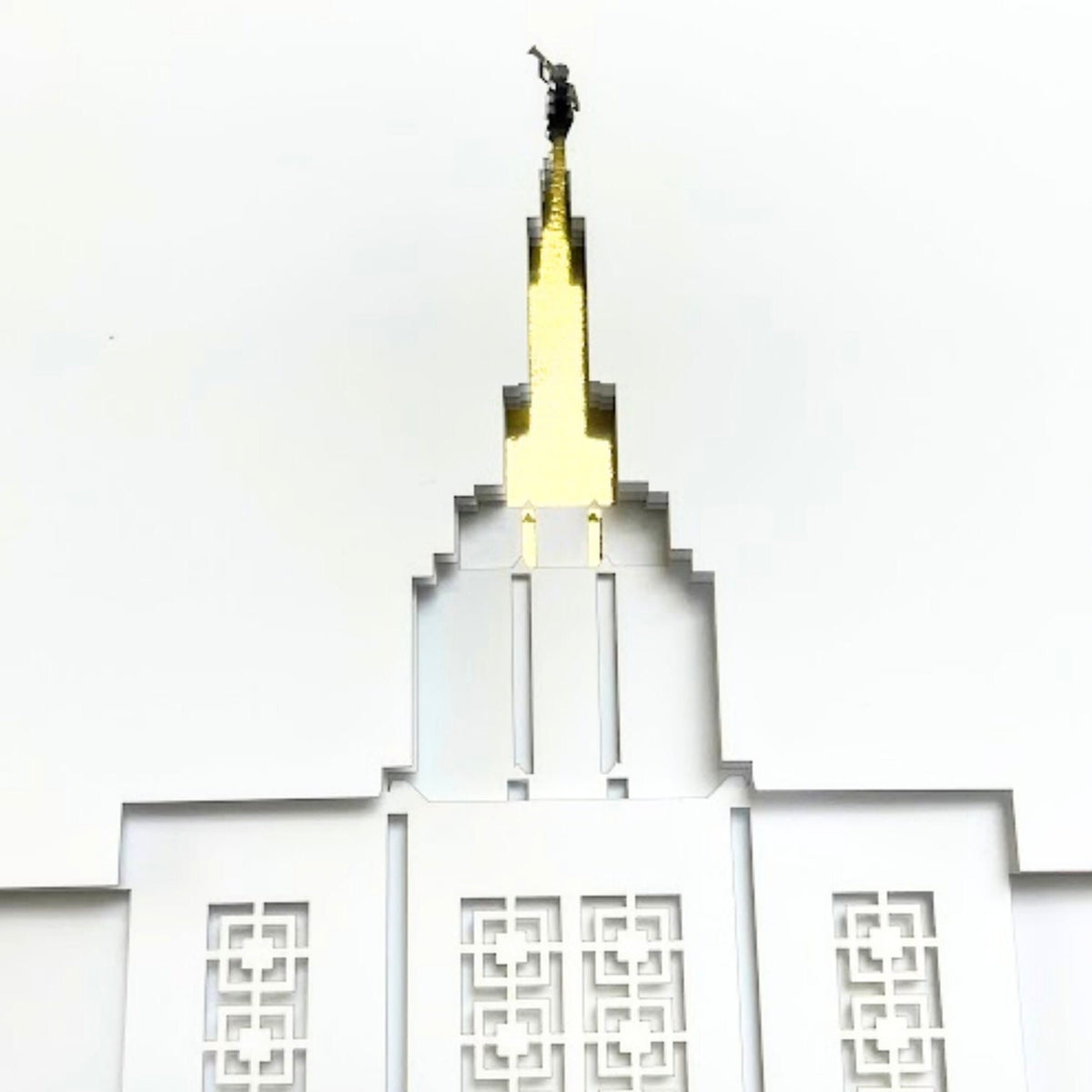 A close-up of a white architectural model or cutout featuring a golden spire and a small dark statue on top, set against a plain white background.