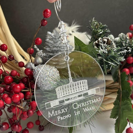 A round clear acrylic Christmas ornament featuring a white engraved illustration of a war memorial building and the text 'MERRY CHRISTMAS Provo 1st War', hanging in front of a holiday wreath with red berries and green foliage.