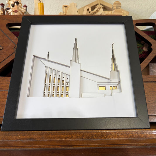 A framed 3D papercut sculpture of the Las Vegas Temple building, displayed on a wooden surface. The sculpture features white layered paper cutouts with gold accents representing windows and architectural details, set against a white background inside a black square frame.