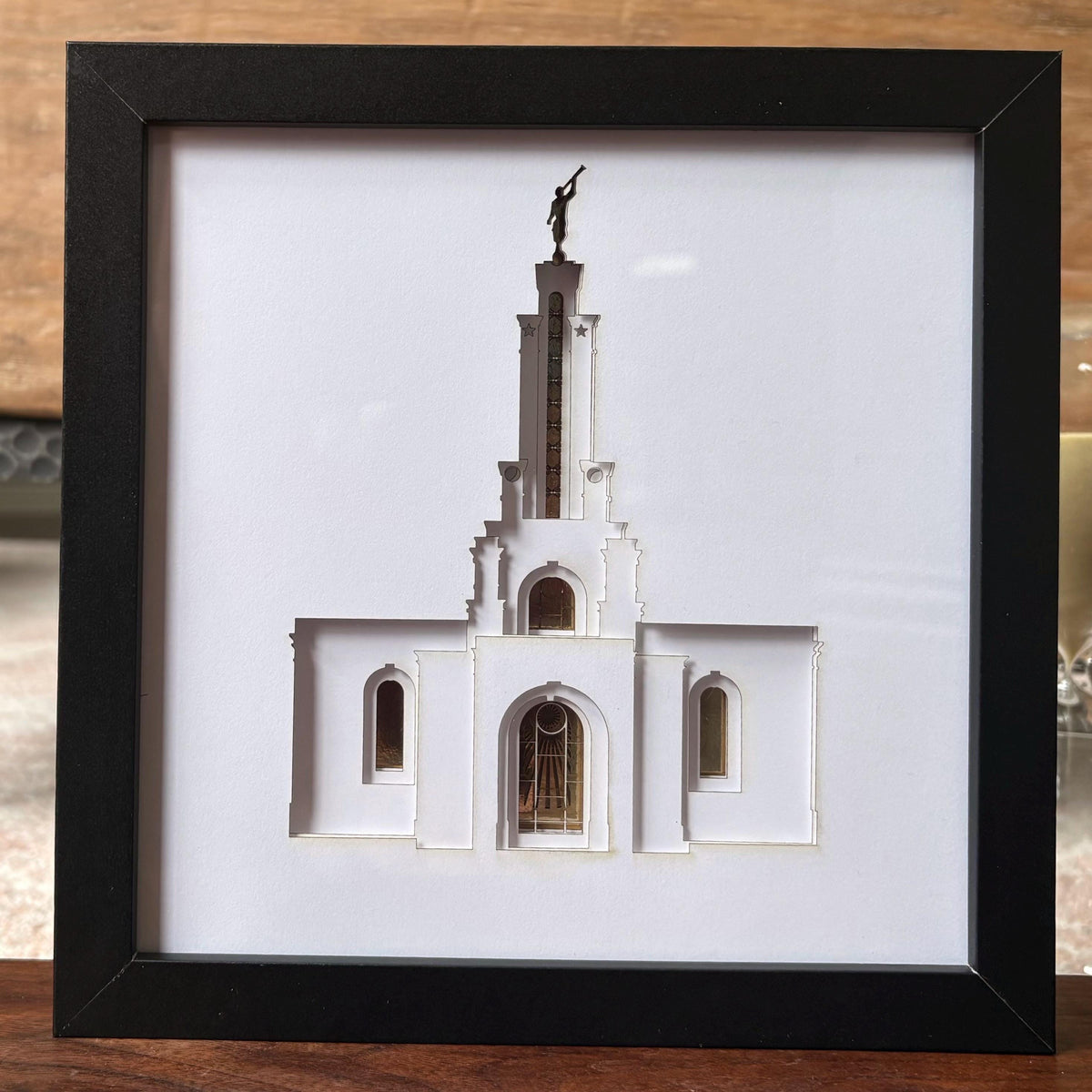 A framed 3D papercut sculpture of the White Temple in Salt Lake City, featuring a white temple structure with a gold-colored stained glass window and a bronze statue on top, set in a black frame. Lubbock Temple Shadow Box Art