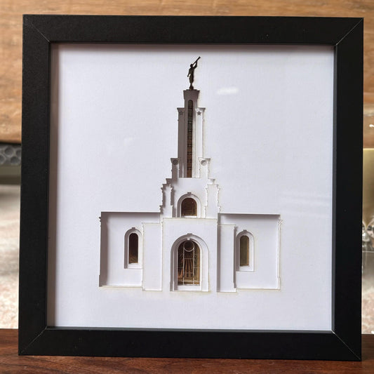 A framed 3D papercut sculpture of the White Temple in Salt Lake City, featuring a white temple structure with a gold-colored stained glass window and a bronze statue on top, set in a black frame. Lubbock Temple Shadow Box Art