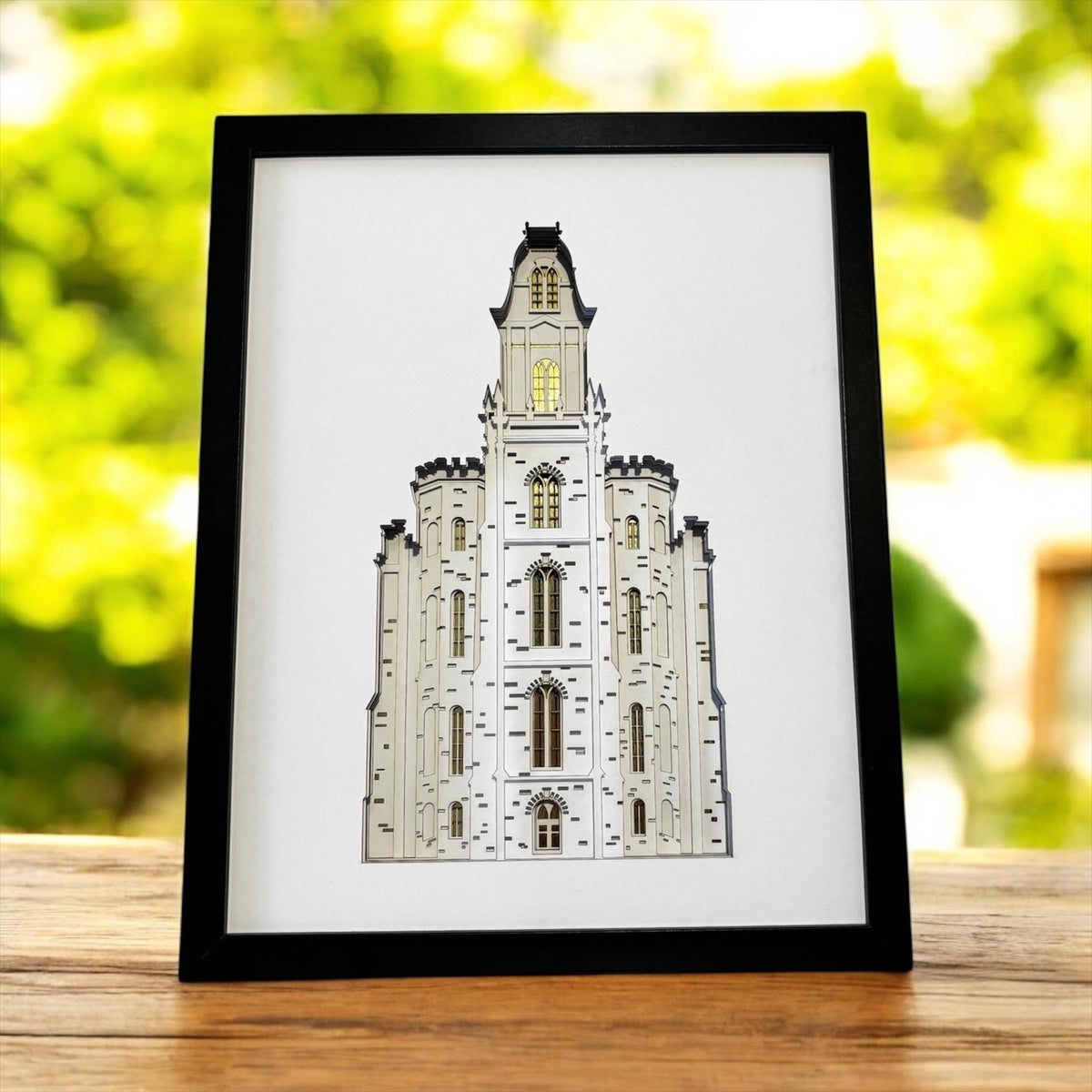 A framed art print of the Salt Lake Temple in Salt Lake City, Utah, featuring a detailed white and gray architectural illustration with a gold-toned clock window. The print is housed in a black frame and is displayed on a wooden surface with a blurred green outdoor background. Manti Temple Art Shadow Box