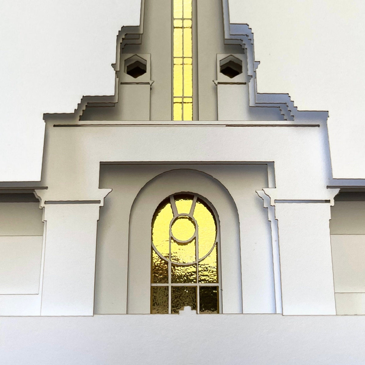 A close-up of a white architectural or decorative wall panel with stepped geometric edges and two arched window sections, one with a yellow-gold stained-glass style center panel. Mt. Timpanogos Temple Art Shadow Box