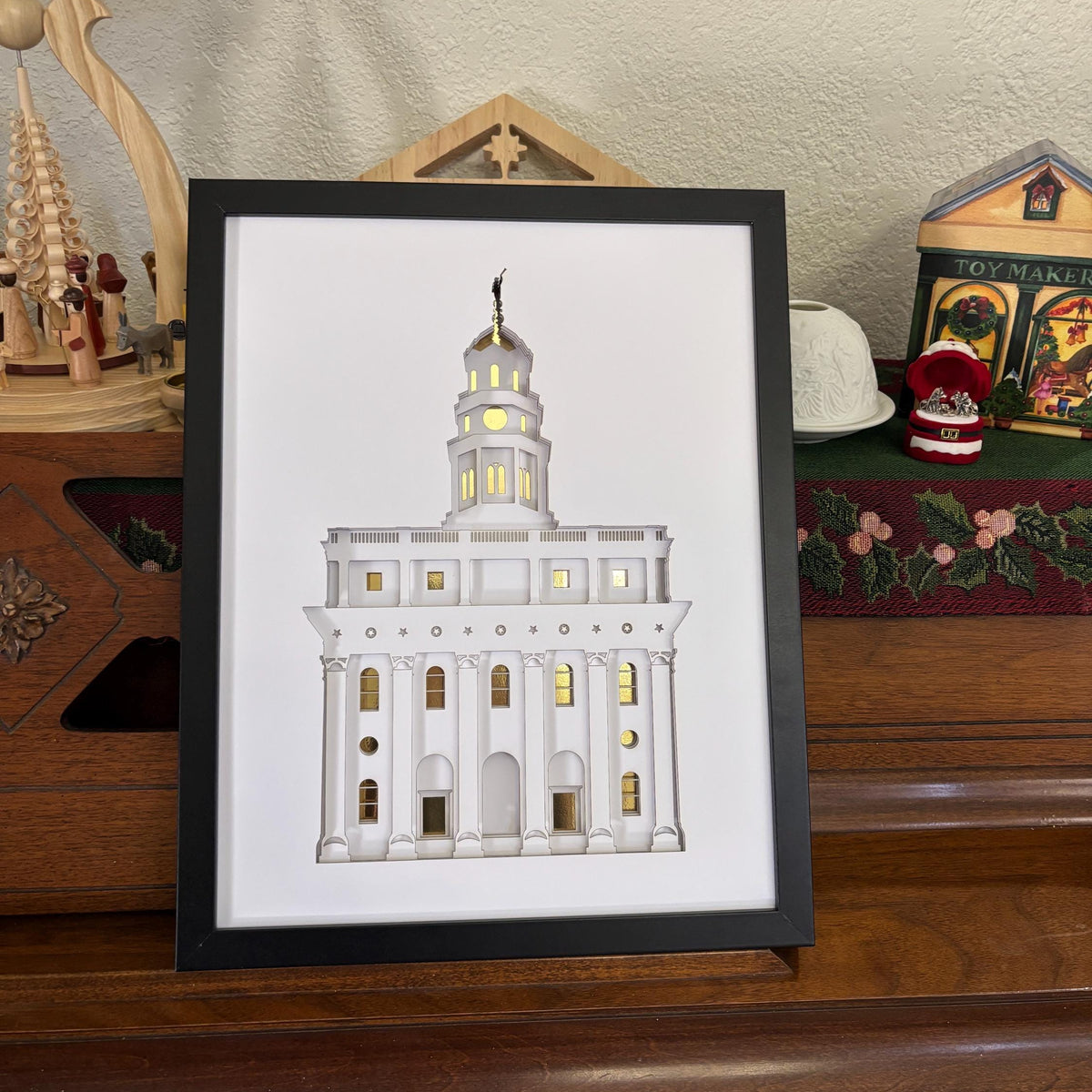 A framed art print featuring a white architectural illustration of a temple building with gold window highlights, displayed on a wooden surface in a home setting.