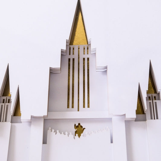 A white paper-cut style decorative structure with multiple pointed spires and vertical gold foil accents, photographed against a plain white background. Oakland Temple Art Shadow Box