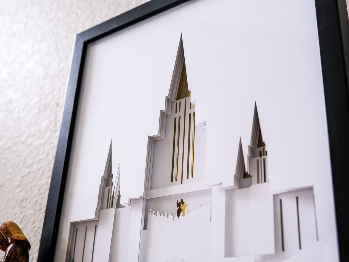 A framed 3D papercut wall art piece depicting a white temple with gold accents, shown hanging on a textured beige wall.
