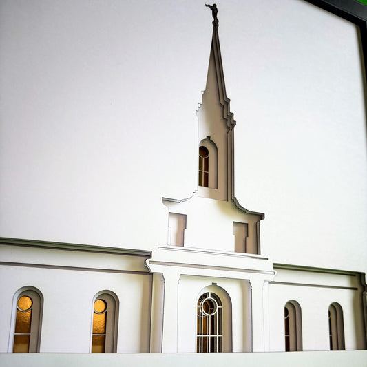 A close-up of a white architectural model or miniature depicting a church facade with a tall pointed steeple, arched windows, and warm glowing window light against a plain light background.