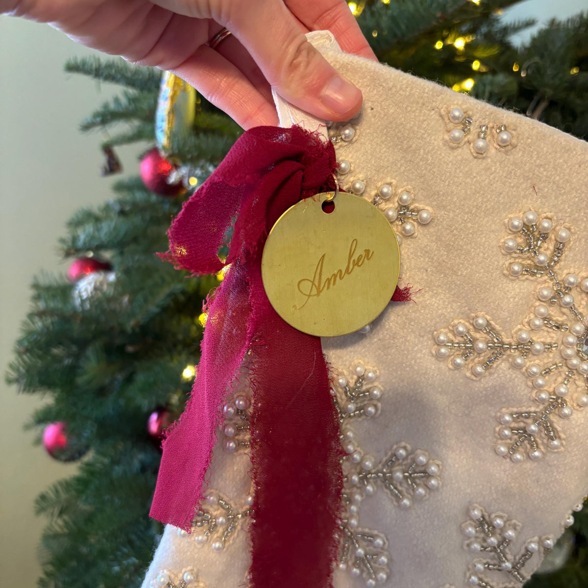 A hand holds a white Christmas stocking decorated with silver bead snowflakes and a dark red ribbon. A round gold tag on the ribbon is embroidered with the name 'Amber'.