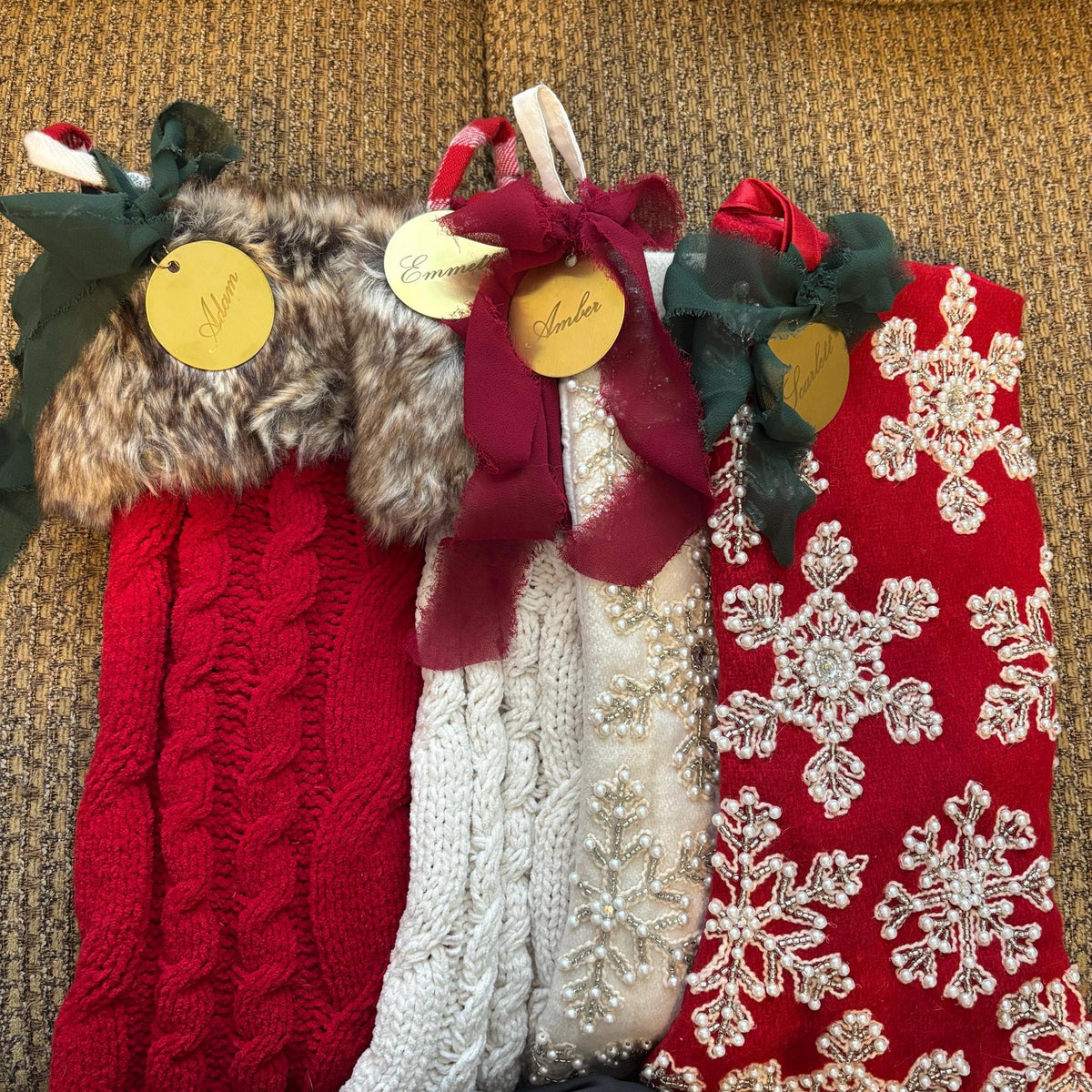 Three Christmas stockings displayed side-by-side on a textured mustard-yellow couch. The left stocking is red cable-knit with a faux-fur trim and green bow.