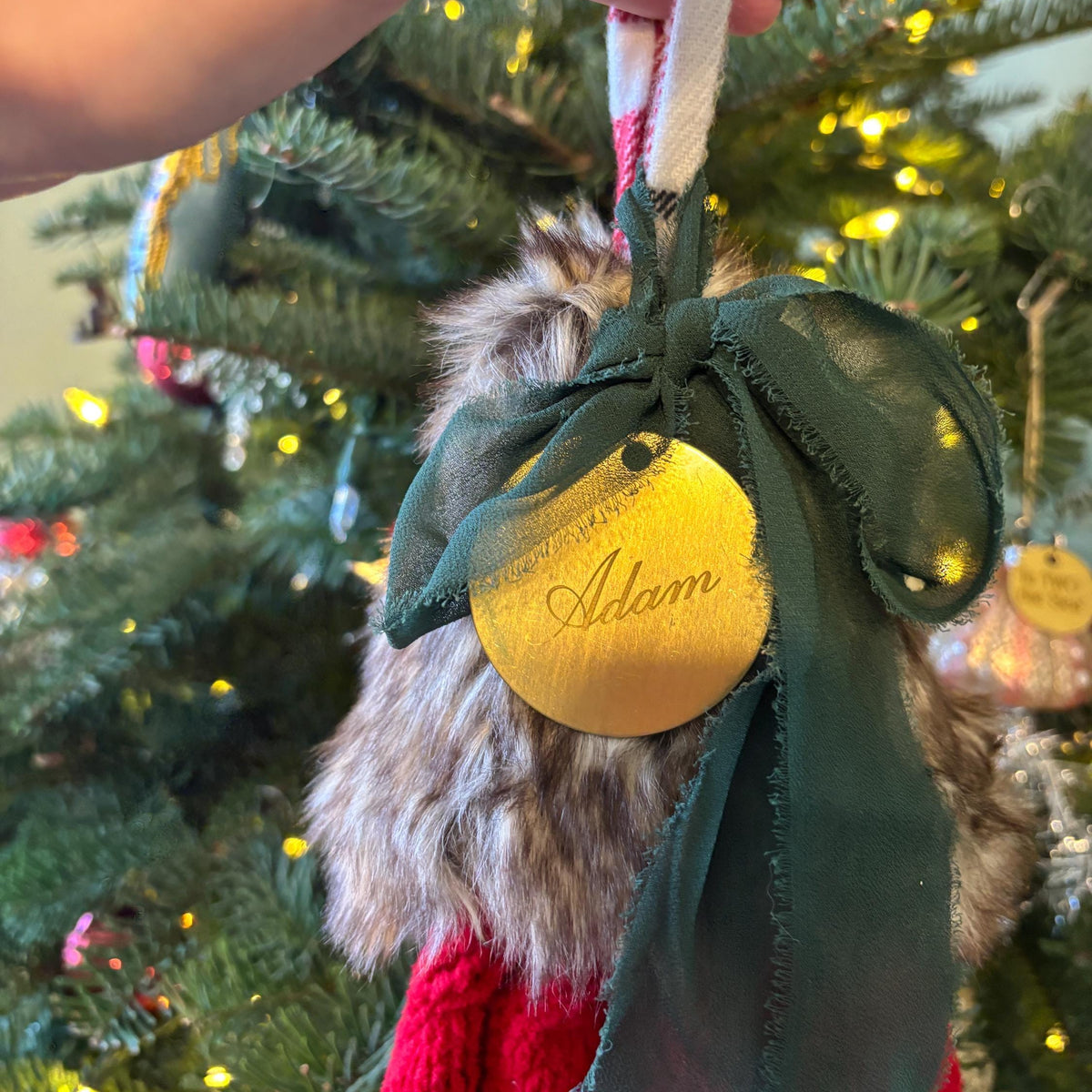 A close-up of a hand hanging a personalized Christmas ornament on a decorated Christmas tree. The ornament is a round gold tag with the name 'Adam' in cursive, tied with a dark green ribbon and a faux fur accent.