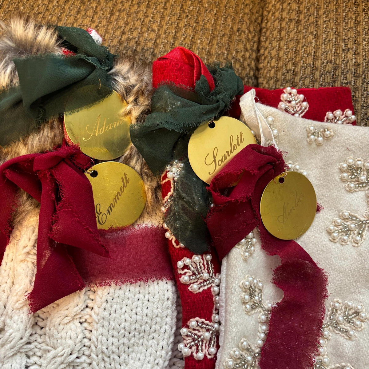 Three Christmas-themed knit accessories laid flat on a textured tan couch: a white cable-knit stocking with a burgundy ribbon and gold tag, a red-and-white patterned stocking with a dark green ribbon and gold tag, and a white knit bootie with faux fur trim, white floral embroidery, and a burgundy ribbon with a gold tag.