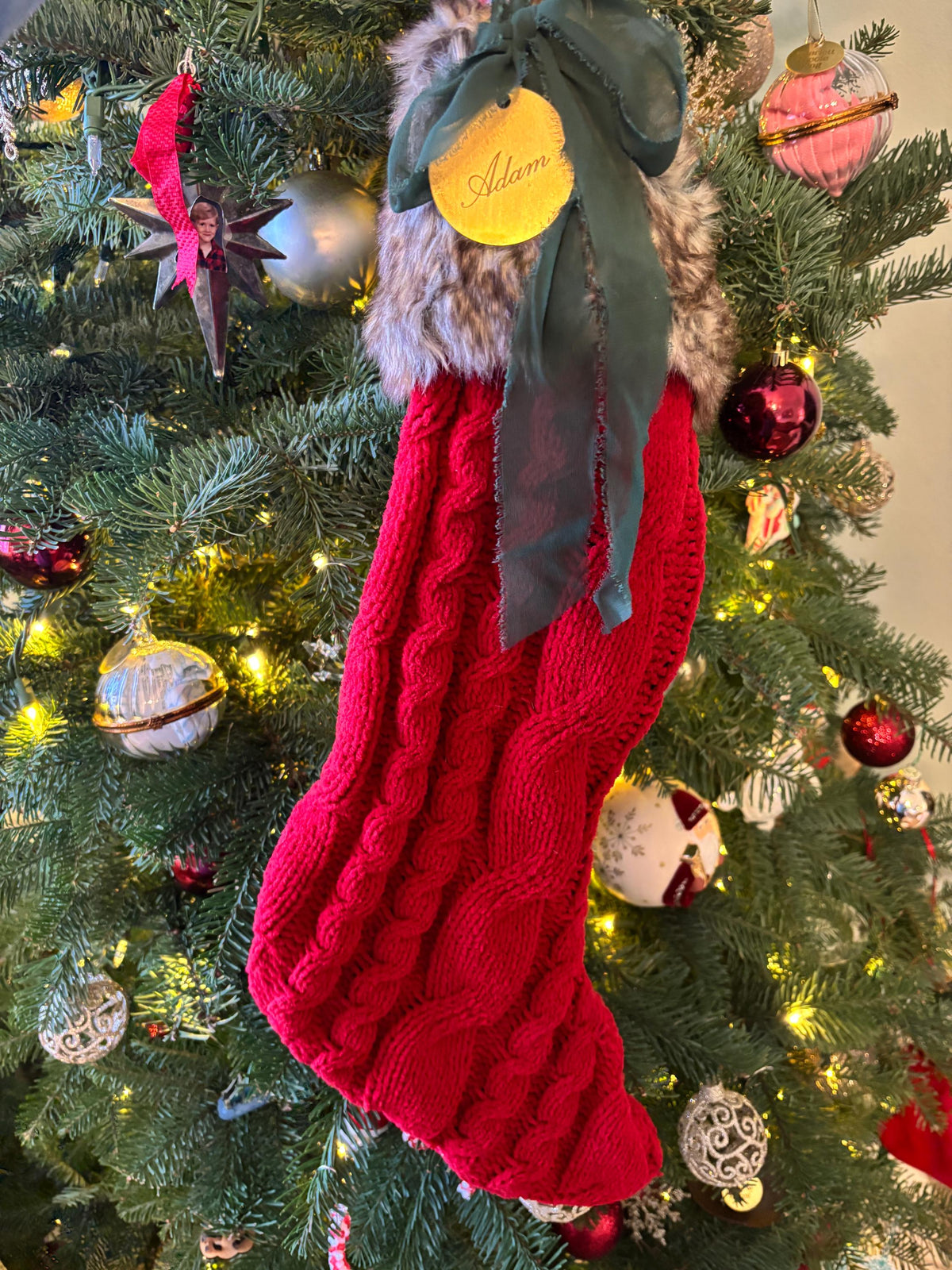 A bright red cable-knit Christmas stocking with a faux fur trim and a dark green ribbon bow, hanging on a decorated Christmas tree. A round yellow tag on the bow reads 'Adore'.