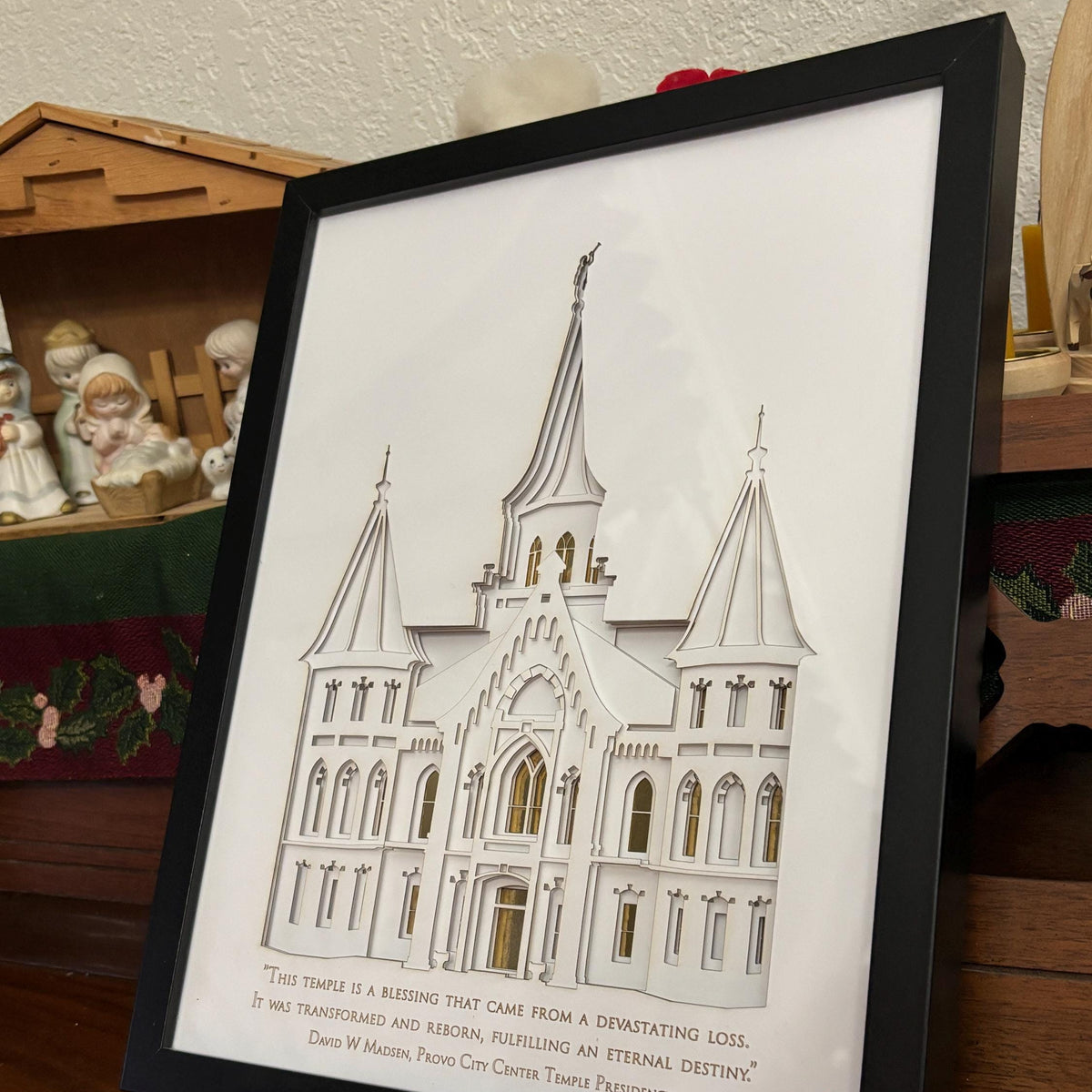 A framed art print featuring a white architectural rendering of a cathedral with a gold-toned inscription at the bottom, displayed on a wooden shelf. Provo City Center Temple Art