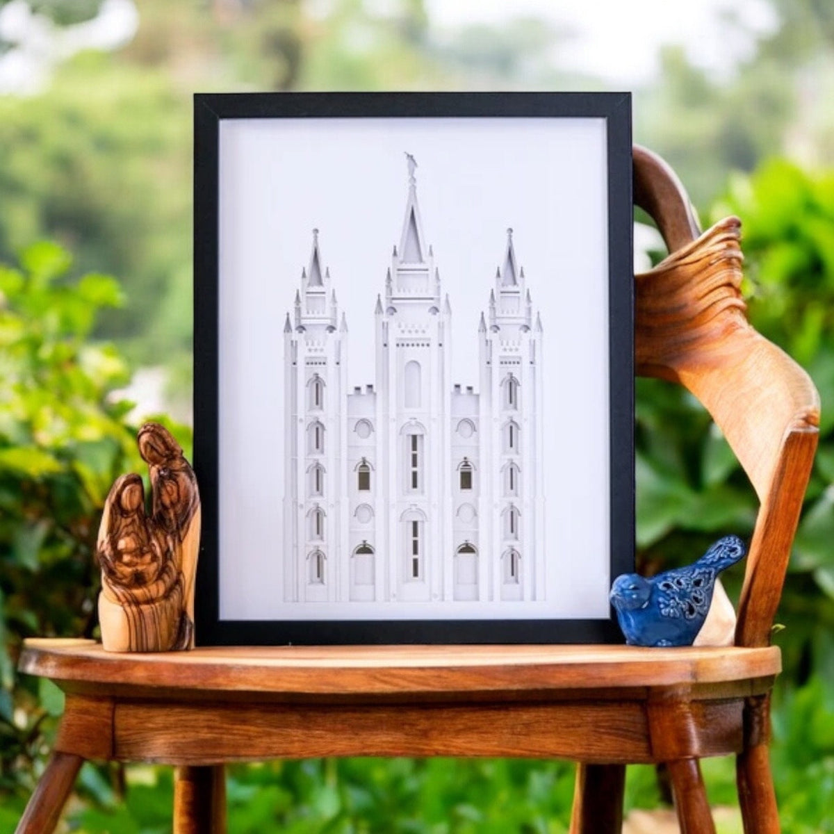A framed black-and-white architectural art print of a cathedral, displayed on a wooden chair outdoors with green foliage in the background.