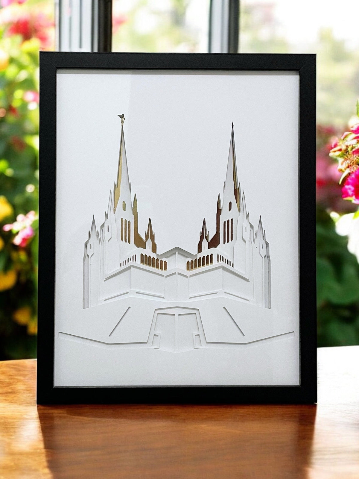 A framed laser-cut architectural art print of the Salt Lake Temple, featuring white layered cutouts with gold foil highlights inside a black frame, displayed on a wooden table with a blurred garden background. San Diego Temple Art Shadow Box