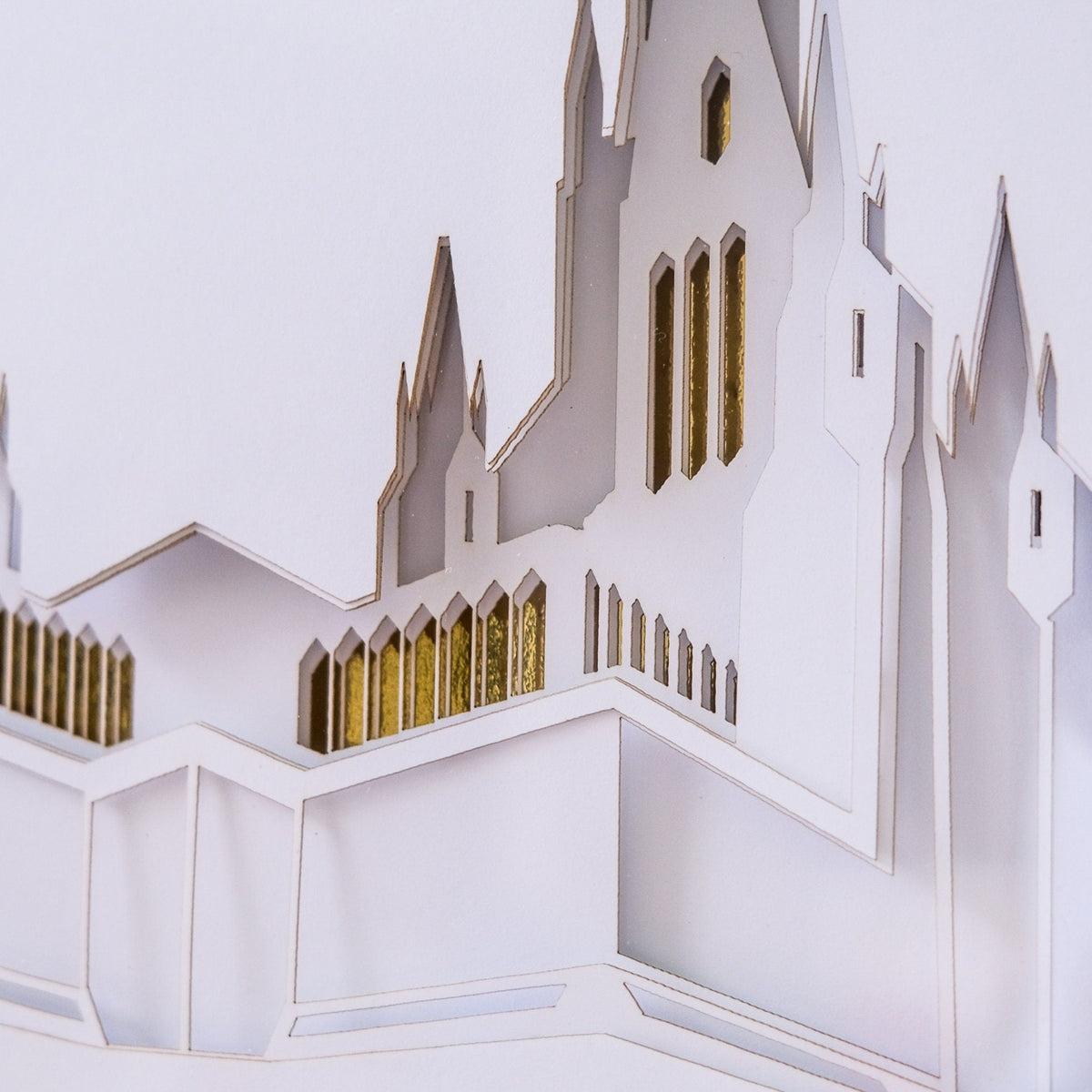 A close-up of a white layered paper cutout model of a castle or cathedral with gold-colored window accents, photographed against a plain white background. San Diego Temple Art Shadow Box