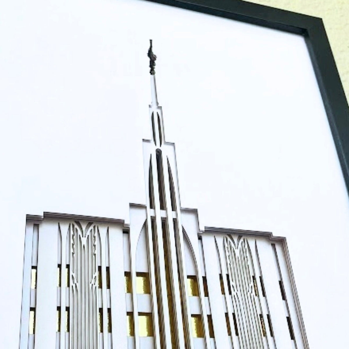 Framed laser-cut wall art featuring a stylized Art Deco temple spire design in white and silver tones with gold window accents, set in a black frame.