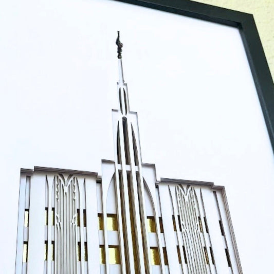 Framed laser-cut wall art featuring a stylized Art Deco temple spire design in white and silver tones with gold window accents, set in a black frame.
