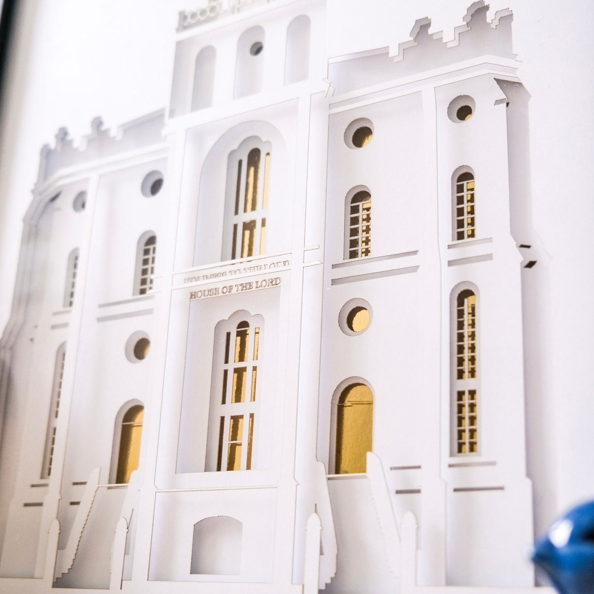 A close-up of a white architectural model or miniature building facade with gold-toned window frames and doors, photographed against a white background. St. George Temple Art Shadow Box