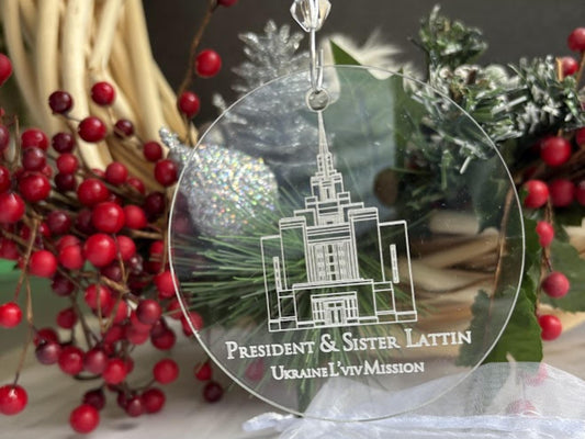 A round clear acrylic Christmas ornament featuring a white line-art engraving of a temple and the text 'PRESIDENT & SISTER LATTIN' and 'UKRAINE LVIV MISSION.' The ornament is shown hanging in a holiday setting with red berries, greenery, and a silver ornament in the background.