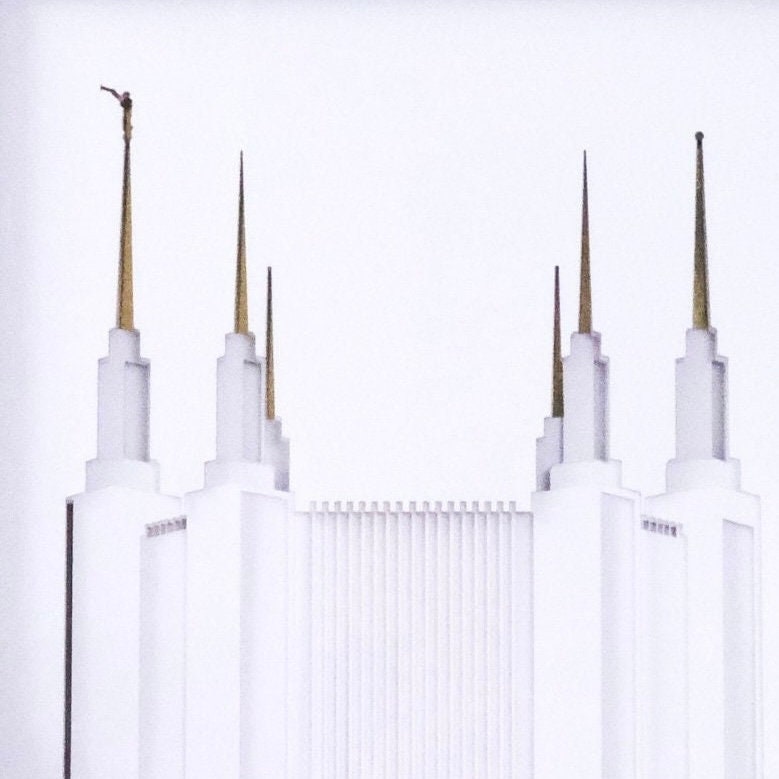 A minimalist architectural photograph of a white temple with tall gold spires against a very light gray or off-white background.
