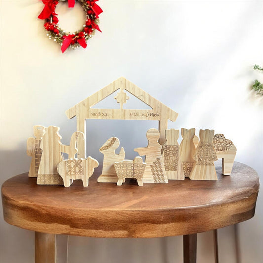A wooden nativity scene set displayed on a round wooden table against a white wall, featuring a stable structure and multiple small wooden figures with engraved patterns and text.